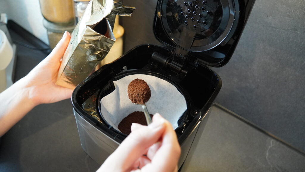 Eine Person hält einen Löffel mit Kaffeepulver über einen Filter in einer schwarzen Kaffeemaschine.