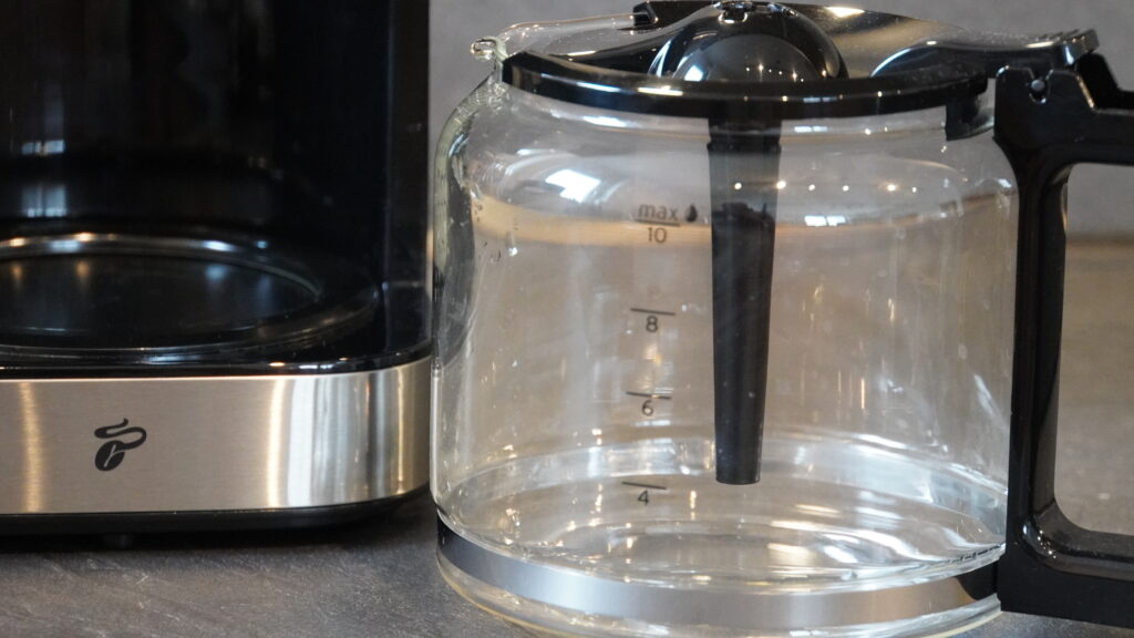 Glaskanne mit Messskala für eine Kaffeemaschine, neben der Maschine platziert, mit schwarzem Griff und Edelstahlakzenten.