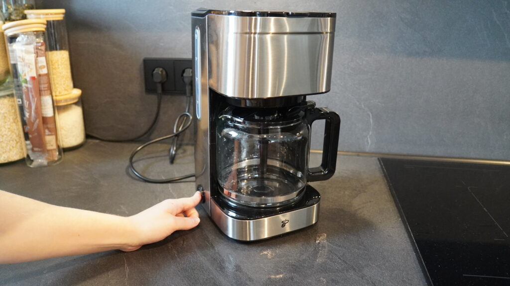 Kaffeemaschine mit Glaskanne und einem Bedienknopf auf der Vorderseite, auf einer dunklen Arbeitsfläche platziert.