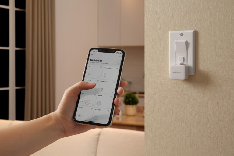Hand hält Smartphone mit SwitchBot App, daneben ein weißer SwitchBot Wiederaufladbarer Bot an der Wand neben einem Schalter, mit Akku‑Fokus