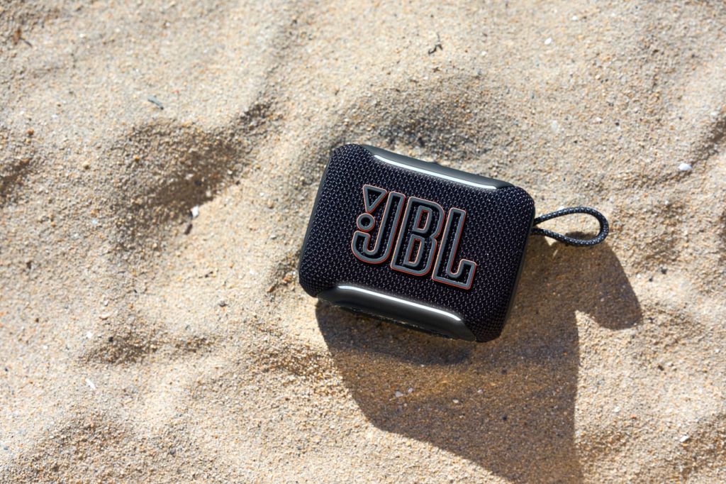 Kleiner, schwarzer JBL Tragbarer Speaker mit texturierter Oberfläche und Schlaufe, liegt auf Sand; kompakt, outdoor-tauglich, zeigt das ikonische JBL-Logo