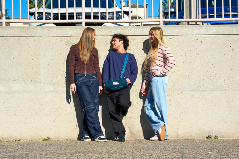 Drei Jugendliche stehen vor einer Betonmauer, einer hält einen blauen JBL-Beutel mit Logo, alle tragen jeans und lächeln; Hintergrund mit Autos