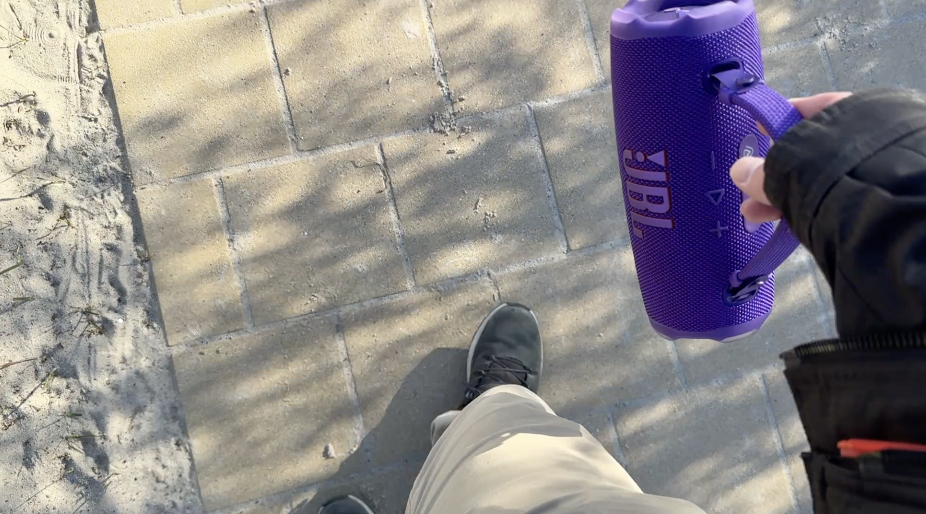 Lila tragbarer Lautsprecher mit gepunkteter Oberfläche und Griff wird auf einem grauen Pflasterweg gehalten; seitlich sichtbare Bedienelemente.