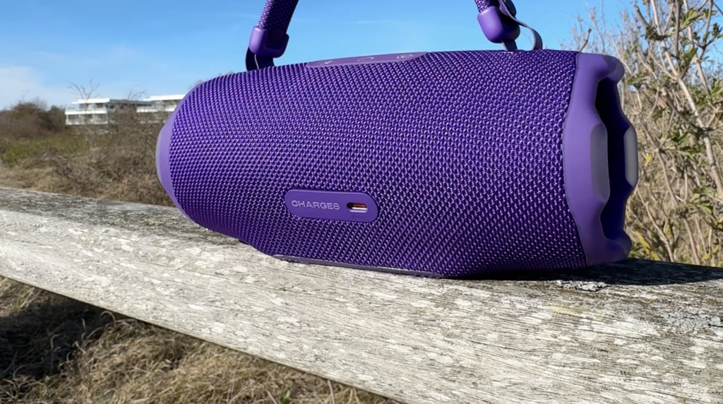 Lila JBL Charge 6 mit abnehmbarem Tragegriff, Text „CHARGE6“ und grobem Gewebemuster auf der Rückseite, draußen bei Tageslicht.