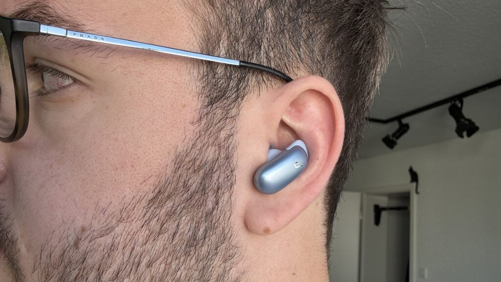 Im Ohr sitzt ein blauer Soundcore Liberty 5 Pro In-Ear-Kopfhörer von Anker, während eine Brille von Prada das Gesichtstreffen begleitet.