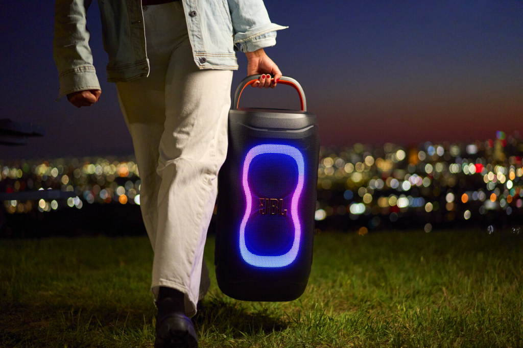 Eine tragbare Bluetooth-Lautsprecherbox mit blauem LED-Licht und lila Leuchtelementen wird nachts im Park getragen, während Lichter der Stadt verschwimmen.