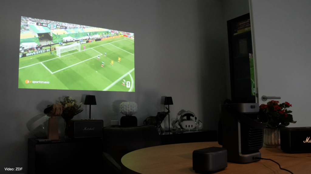 Im Wohnzimmer projiziert der Soundcore Nebula P1 ein ZDF-Fußballspiel an die Wand, während der mobile Beamer neben dem abnehmbaren Lautsprecher steht.