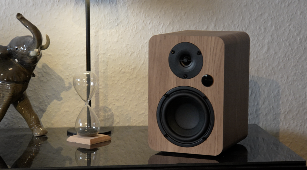 Der kompakte Vulkkano A4 ARC MKII zeigt eine Holzoptik mit schwarzer Front, zwei Treibern und steht neben einer Sanduhr auf einer Glasfläche.