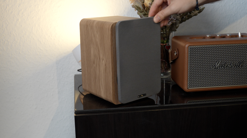 Eine Hand nimmt die graue Frontabdeckung der Vulkkano A4 ARC MKII von einem Lautsprecher in Holzoptik, neben einem braunen Marshall-Boxenradio.