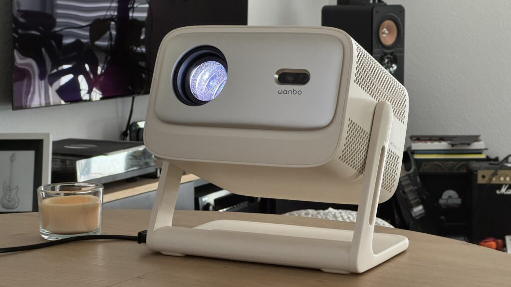 Der helle Wanbo Vali 1 Pro steht auf einem Tisch vor einem Fernseher und Lautsprechern im Wohnzimmer.