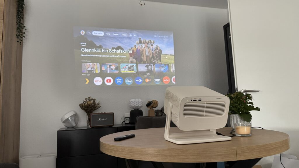 Der beige Wanbo Vali 1 Pro projiziert die Google-TV-Oberfläche mit Prime Video in ein helles Wohnzimmer.