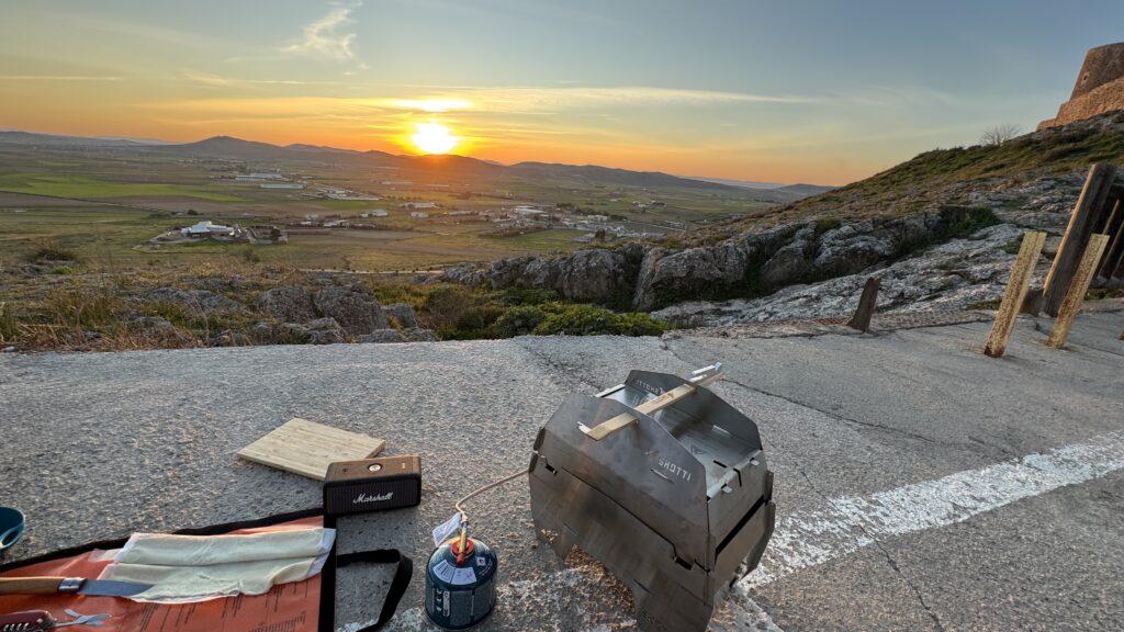 Der SKOTTI Grill steht neben einer Gaskartusche auf einer Steinfläche im Abendlicht über weiter Landschaft.