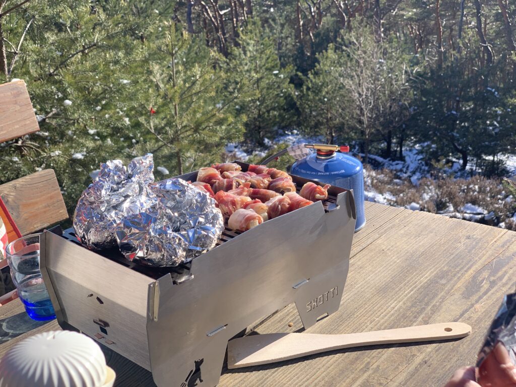 Auf dem SKOTTI Grill liegen Baconröllchen und Folienkartoffeln neben einer blauen Gaskartusche im Wald.
