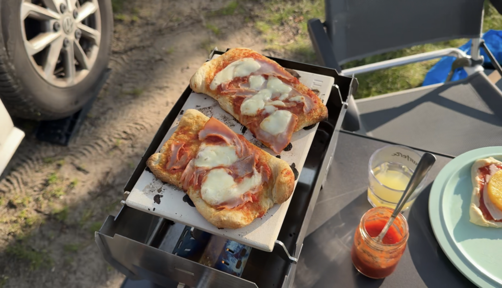 Auf dem SKOTTI Grill backen zwei kleine Pizzen mit Käse und Schinken auf einer Terrasse beim Camping.
