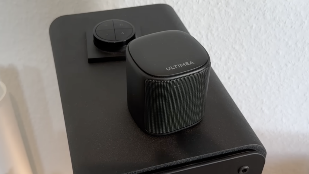 Eine schwarze Ultimea Aura S4T mit kompaktem Lautsprecher und Bedieneinheit steht nah an einer hellen Wand.